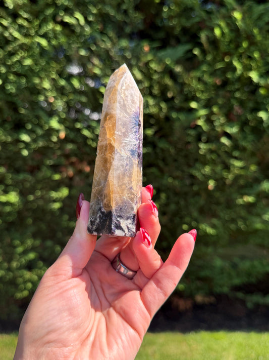 May include: A hand holds a polished crystal tower. The crystal has a yellow, white, and purple color scheme. The base of the crystal is black. The background is a blurred green.