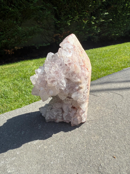 May include: A large, natural crystal formation with a light pink hue and clusters of clear quartz crystals. The crystal sits on a gray surface, with a green lawn and bushes in the background. The crystal's rough texture and unique shape are prominent.