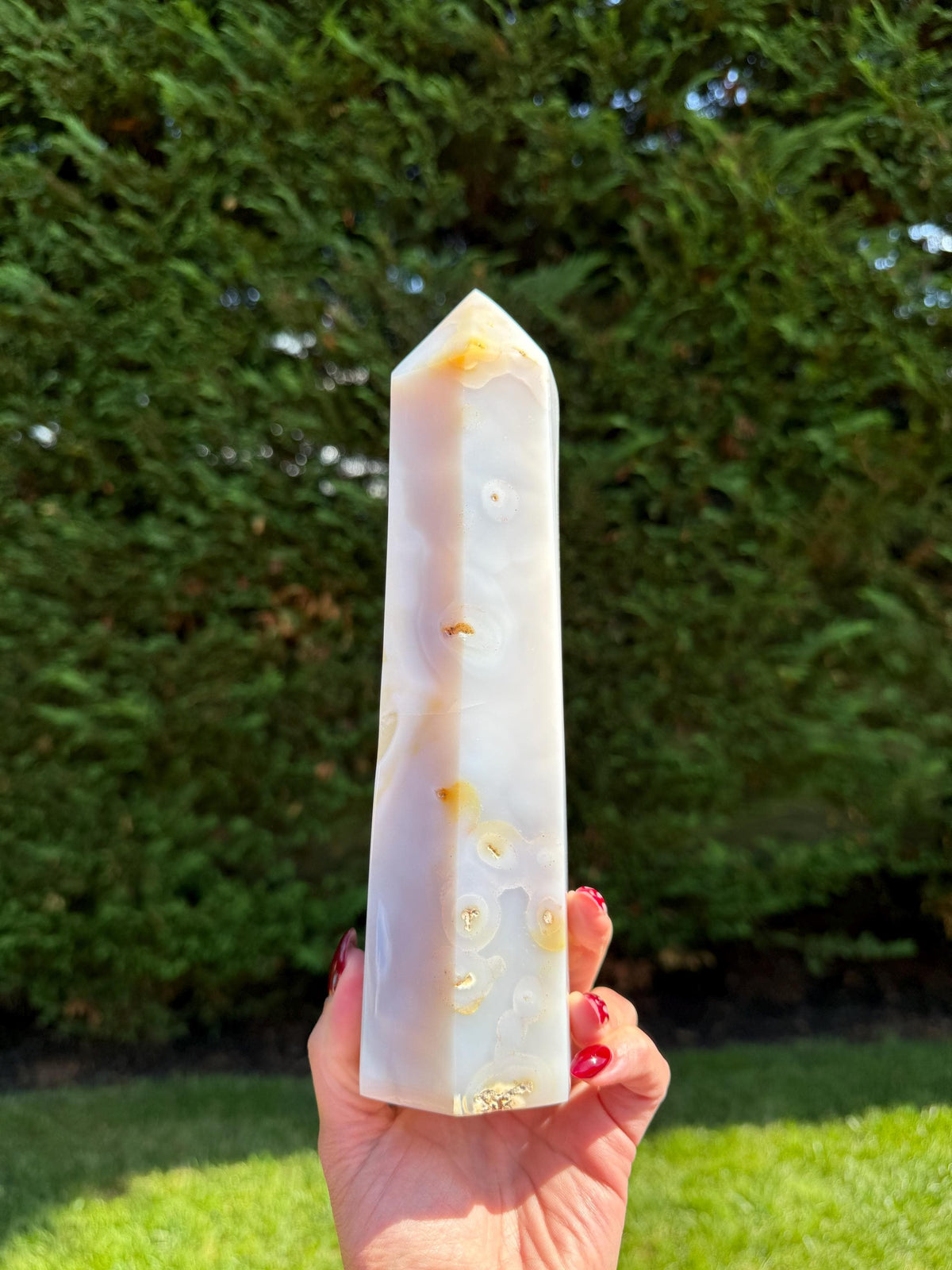May include: A polished agate obelisk, featuring a blend of white, cream, and golden-brown hues. The crystal has a pointed top and a smooth, textured surface. The obelisk is held in a hand with red nail polish, set against a green background.