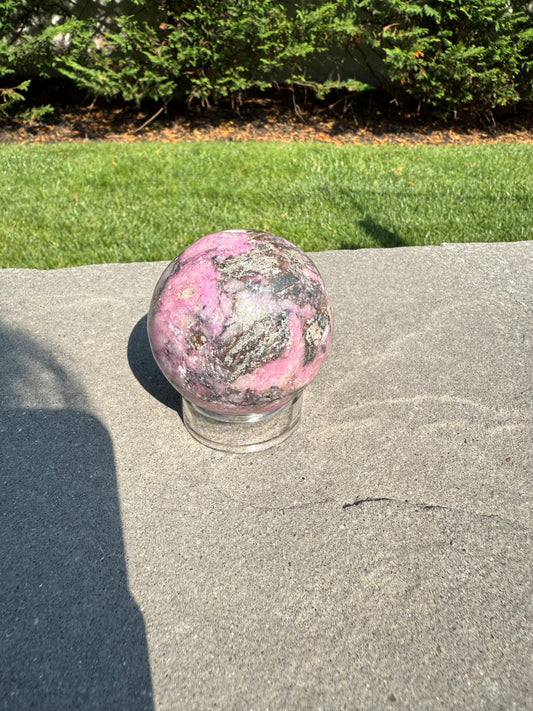 May include: A pink and gray sphere, possibly a crystal ball, sits on a clear glass stand. The sphere has a marbled appearance with shades of pink, gray, and white. The background includes green grass and a hedge.