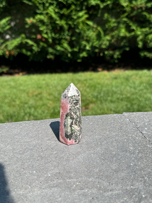 May include: A pink and green crystal point with a pointed tip. The crystal has a natural, rough texture and is a light pink color with green and black inclusions.