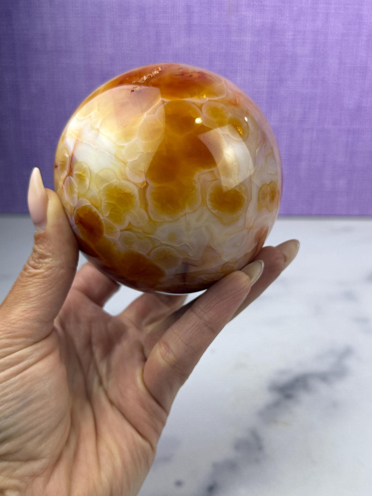 May include: A polished carnelian sphere, showcasing a blend of orange, brown, and white hues. The gemstone's surface has a glossy finish, with a pattern of swirling colors. The sphere is held in a hand against a purple backdrop.