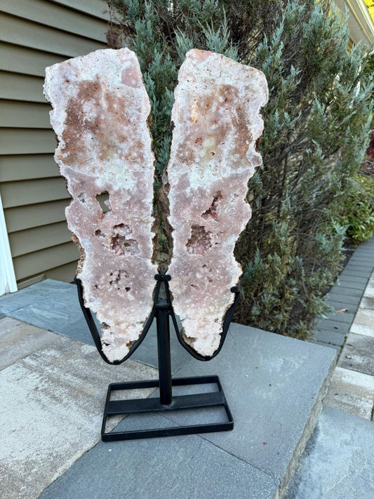 May include: Two large, pink and white geode slices mounted on a black metal stand. The geode has a rough, textured surface with crystal formations and a natural, organic shape. The stand is square at the base and has a curved design.