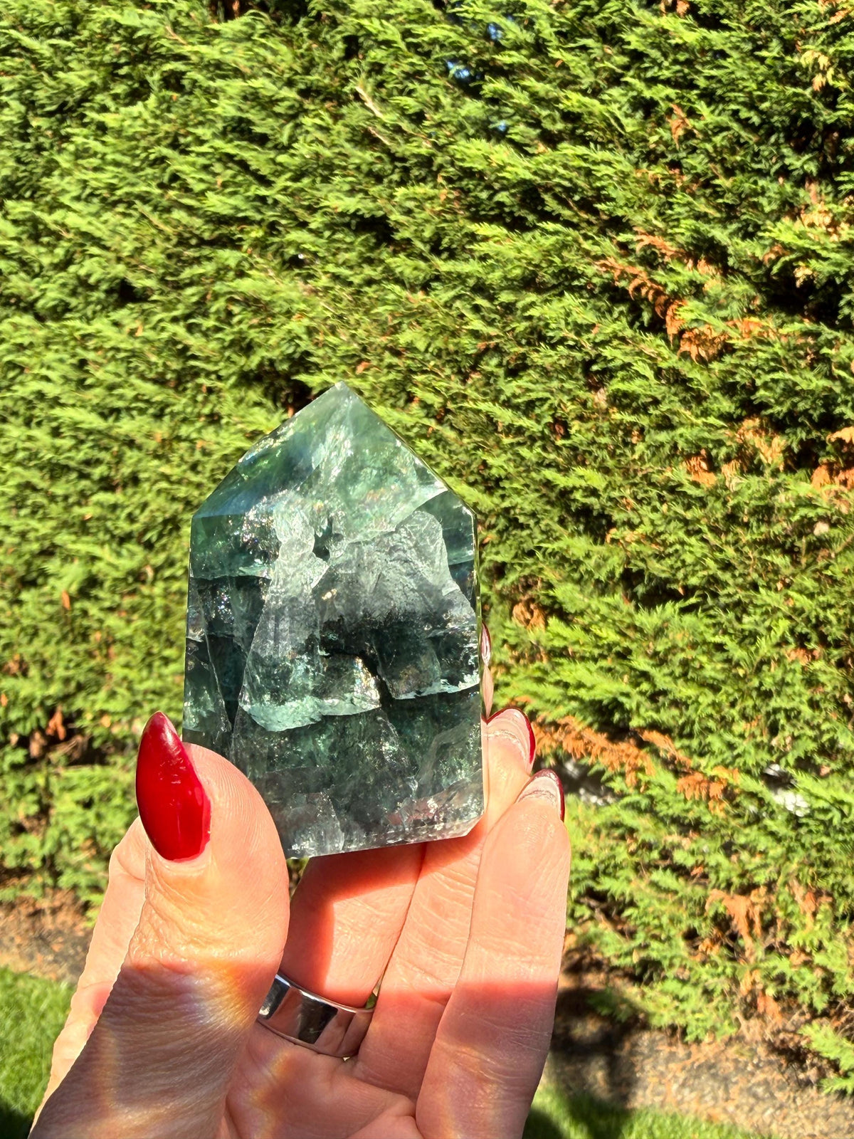 May include: A green and clear fluorite crystal tower with a pointed top. The crystal has a layered appearance with varying shades of green and clear sections. The crystal is held in a hand with red nail polish.