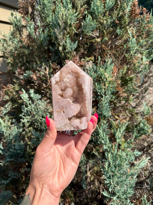 May include: A pink crystal tower with a geode-like interior is held in a hand with red nail polish. The crystal has a natural, rough texture and a light brown hue in the geode cavity. The background features green foliage.