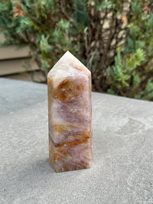 May include: A rectangular crystal obelisk with a pointed top. The stone features a blend of colors, including shades of lavender, cream, and golden brown. The crystal is standing upright on a gray surface, with a blurred green background.