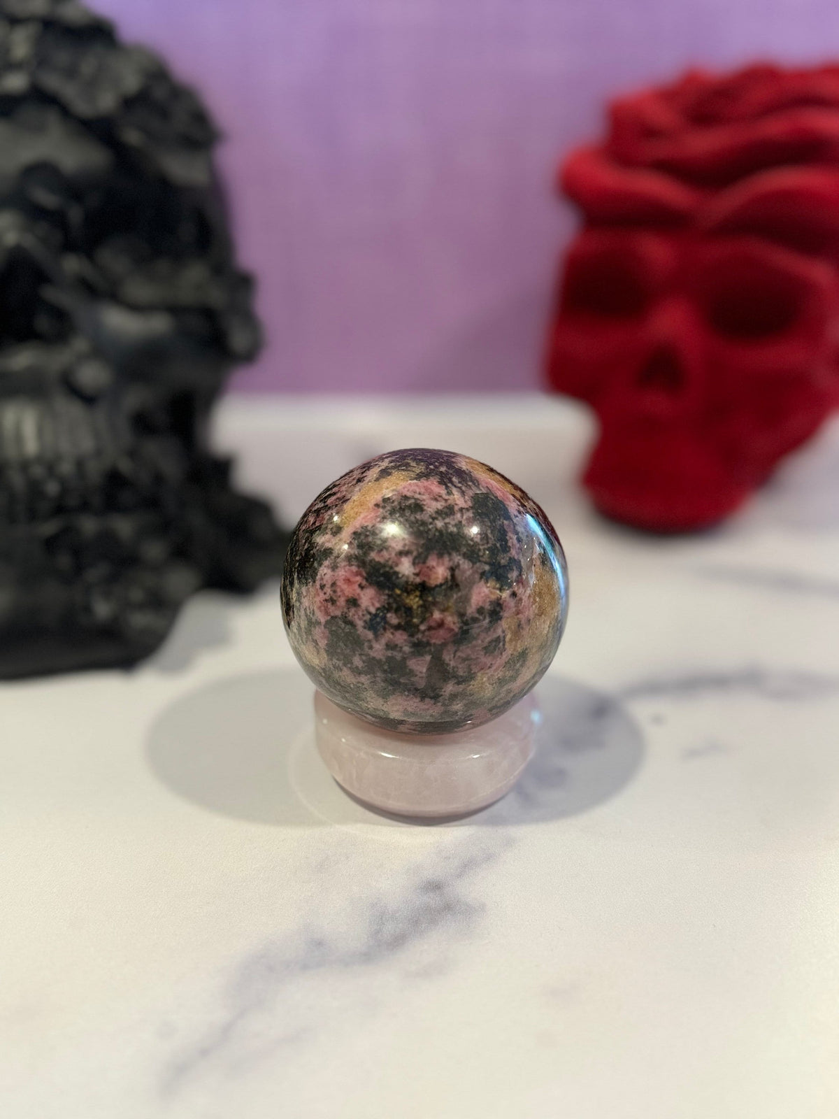 May include: A pink and black sphere of polished rhodonite, a type of gemstone, sits on a white pedestal.
