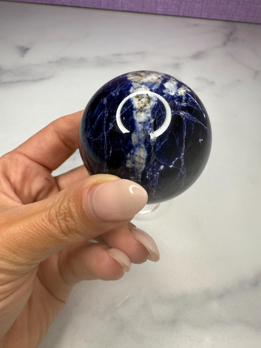 May include: A polished sodalite sphere, showcasing a deep blue hue with white and gray veining. The gemstone ball is held in a clear stand, reflecting the light. This decorative sphere is a unique home decor item.