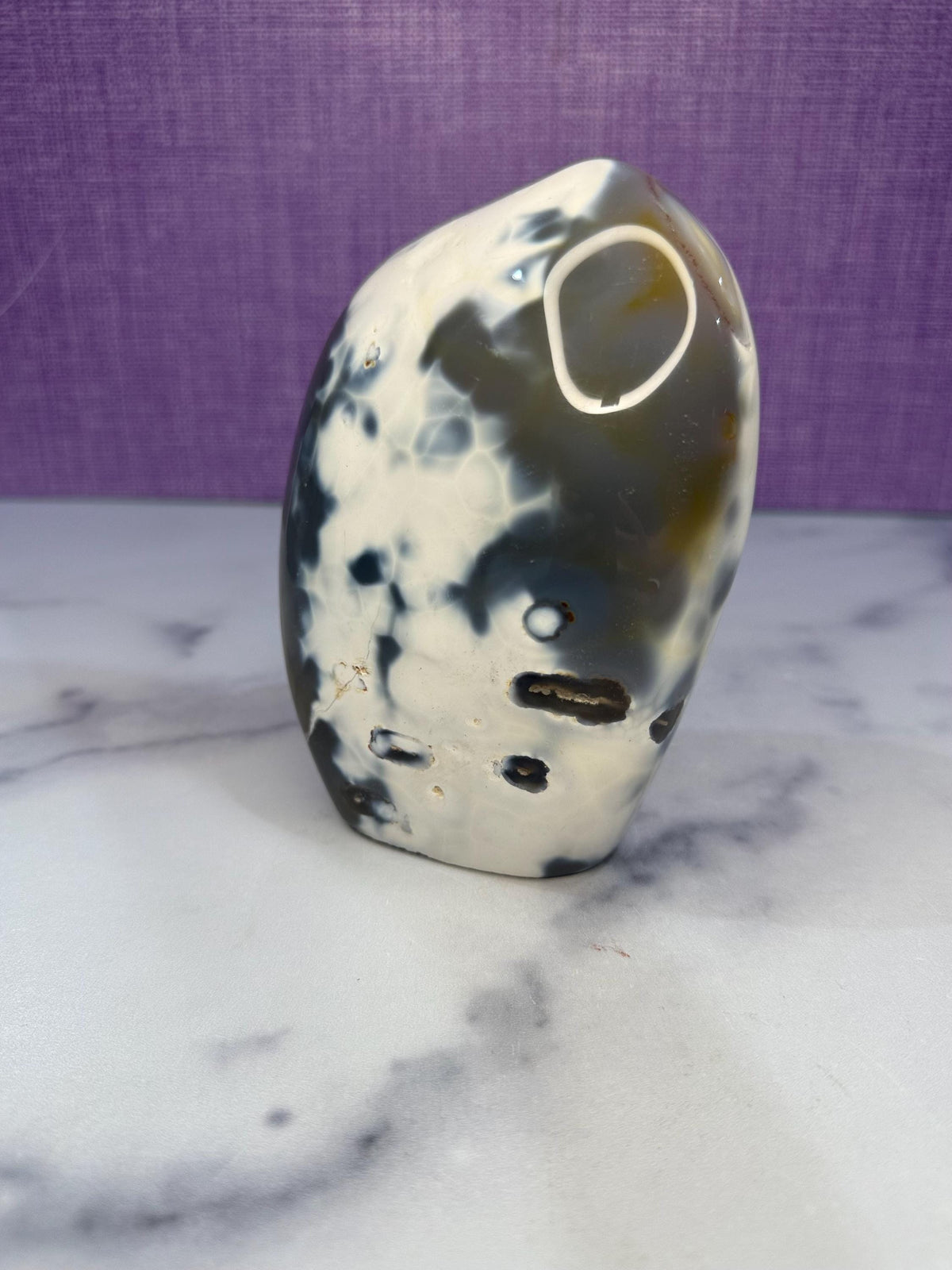 May include: A polished, freeform stone with a mix of colors. The stone features a base of creamy white with dark gray and black patterns. There are also areas of light brown and a large, oval-shaped opening on the side. The stone is displayed on a white marble surface.