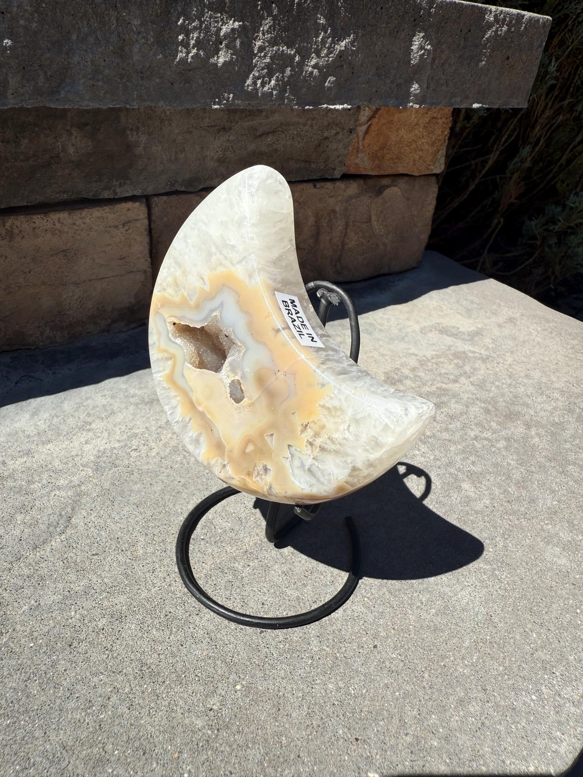May include: A crescent-shaped, polished agate geode with cream, tan, and white coloration, mounted on a black metal stand. The geode has a hollow center with visible crystal formations. A sticker on the geode reads "MADE IN BRAZIL".