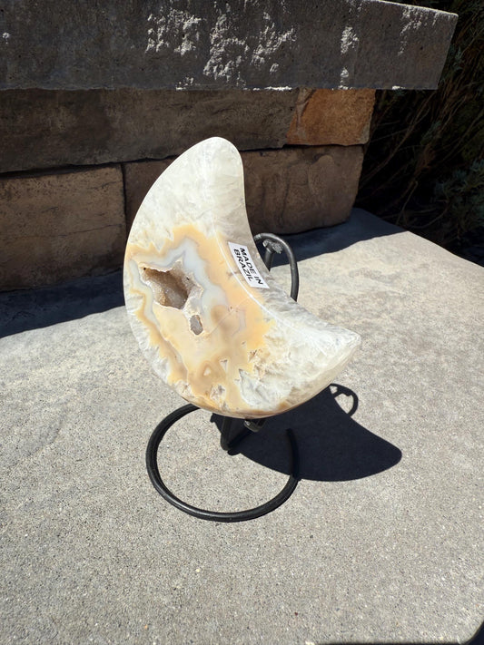 May include: A crescent-shaped, polished agate geode with cream, tan, and white coloration, mounted on a black metal stand. The geode has a hollow center with visible crystal formations. A sticker on the geode reads "MADE IN BRAZIL".