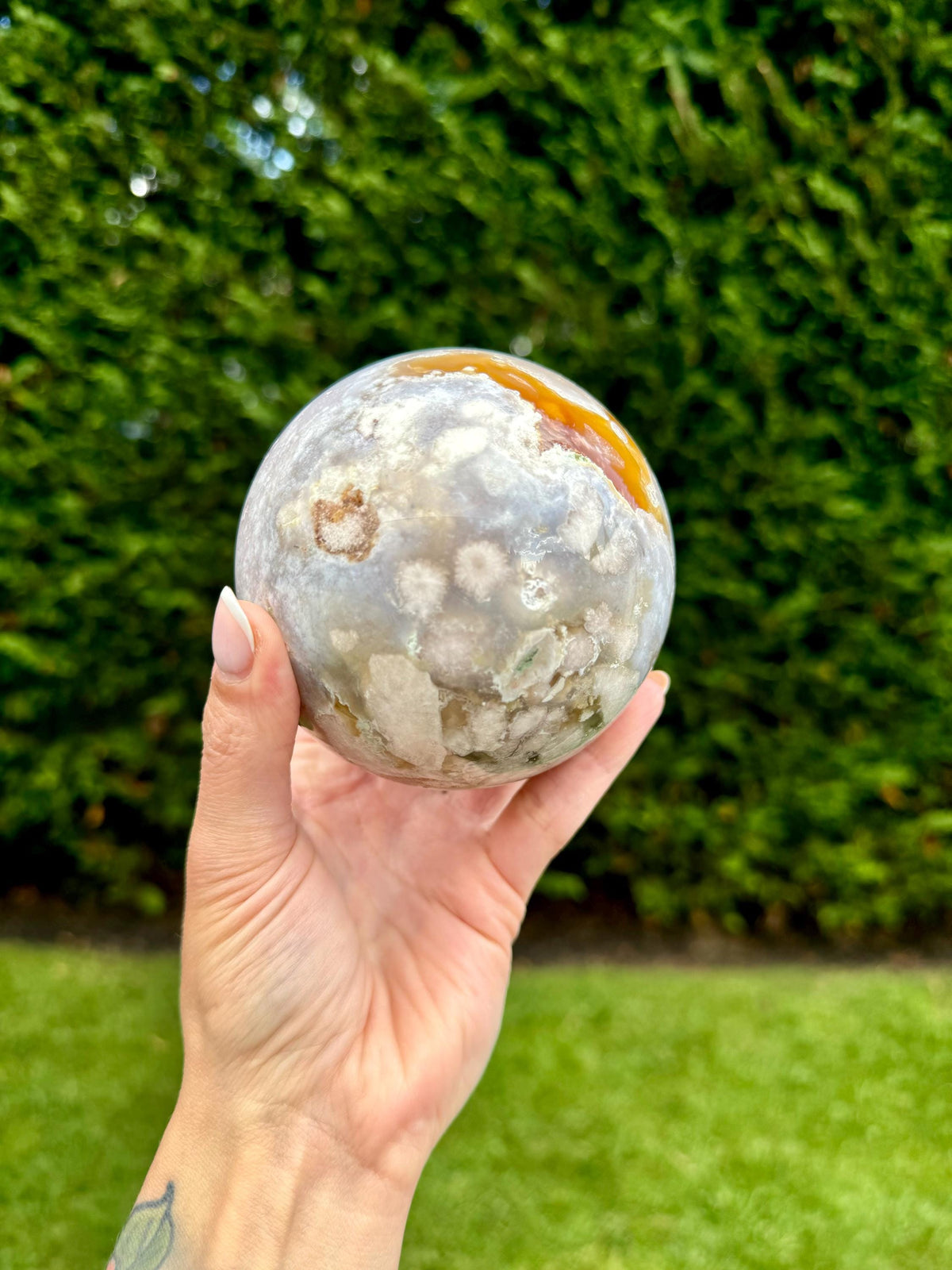 May include: A large, round, gray, white, and orange agate sphere with a variety of patterns and textures. The sphere is being held in a hand.