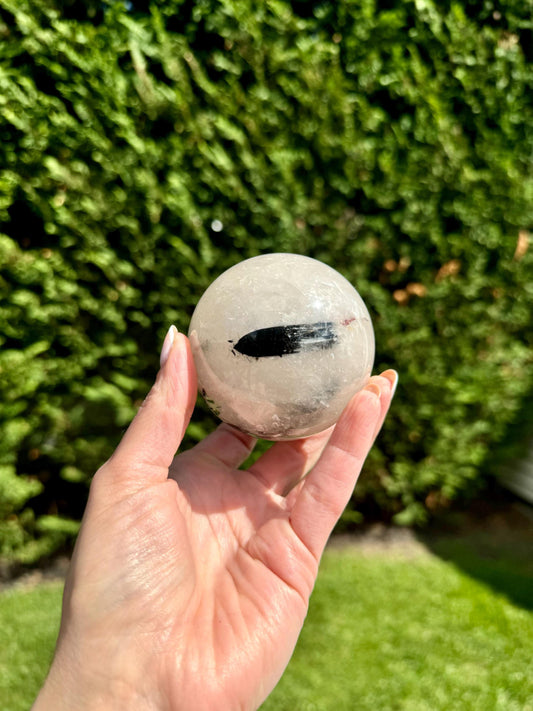 May include: A clear quartz sphere with a black inclusion, polished to a smooth finish. The sphere is held in a hand against a green background.
