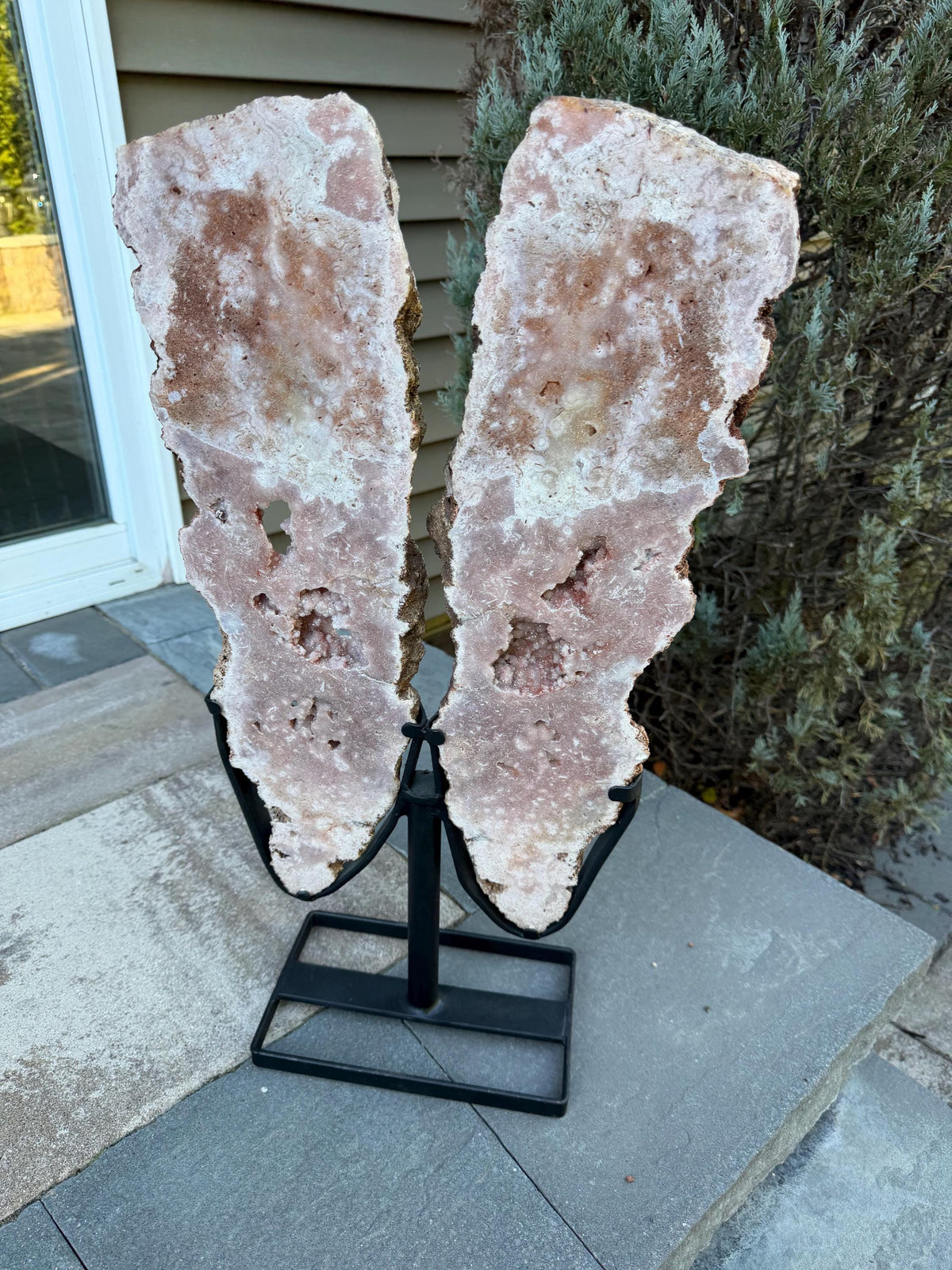 May include: Two large, irregular-shaped pink and white crystal slabs mounted on a black metal stand. The crystal formations have a rough, textured surface with visible cavities and variations in color. The stand has a square base and a central support.